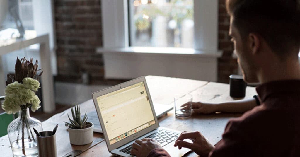 Laptop on a wooden desk with a person working, surrounded by plants and a coffee cup, in a modern office space.