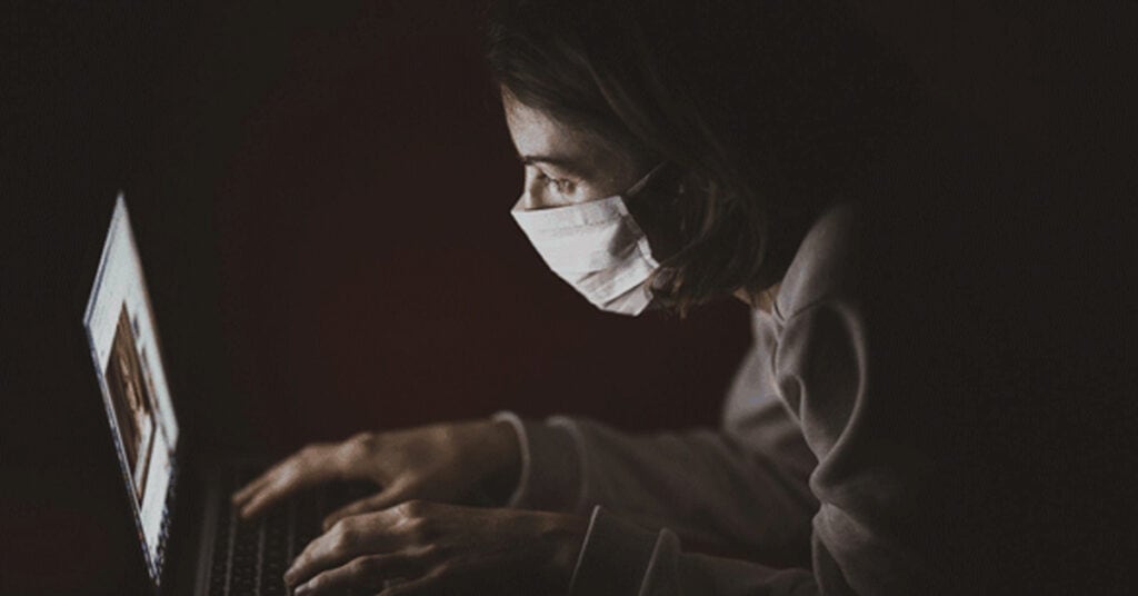 Secure cybersecurity expert working on laptop at night, wearing face mask for health safety, with a focus on digital security and online protection solutions.