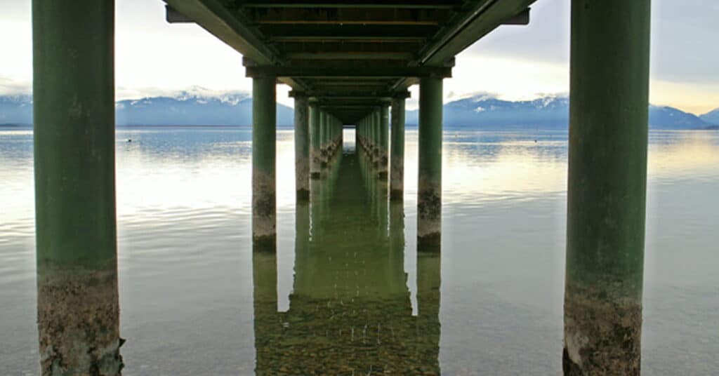 Underwater view of a pier extending into a calm lake with mountains in the background, showcasing infrastructure and nature.