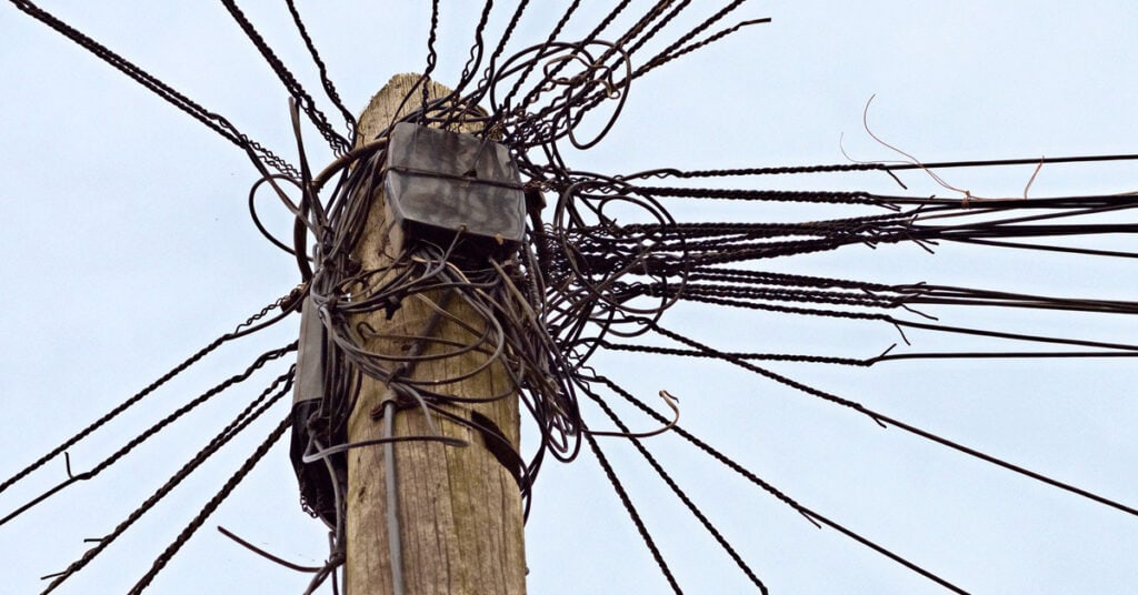Insulated utility pole with tangled electrical wires and an electrical box against a cloudy sky.