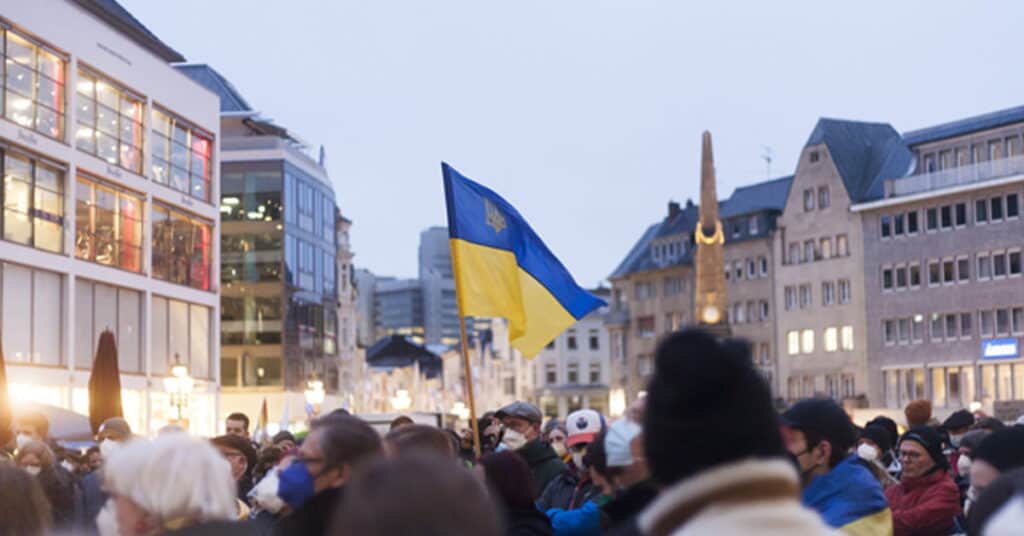Ukrainian flag being held during a public gathering in a city square, demonstrating patriotism and national pride. Crowds of people celebrate amidst modern and historic architecture in an urban environment.